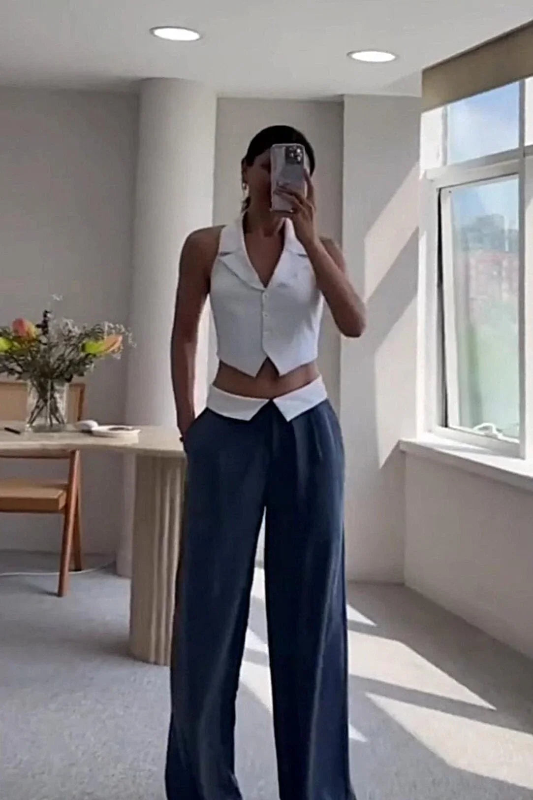 Woman taking mirror selfie wearing white sleeveless vest and navy blue wide-leg pants in bright room