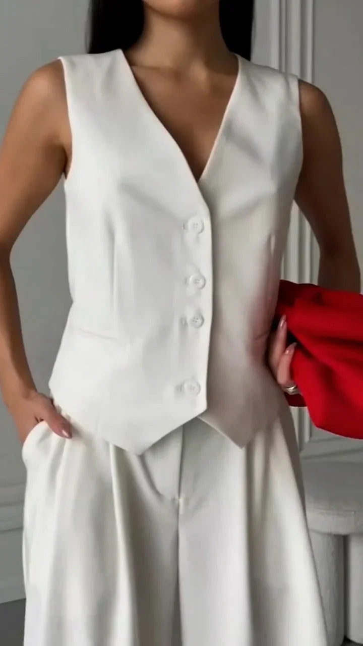 Woman wearing white sleeveless waistcoat and matching pants holding red clutch in a minimalist room