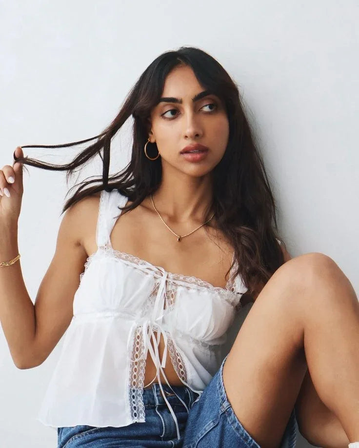 Woman with long dark hair in white lace-trimmed top and denim shorts sitting against white wall