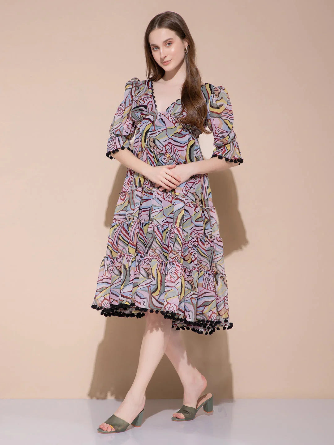 Woman in colorful abstract print midi dress with puff sleeves and black pompom trim posing against peach wall