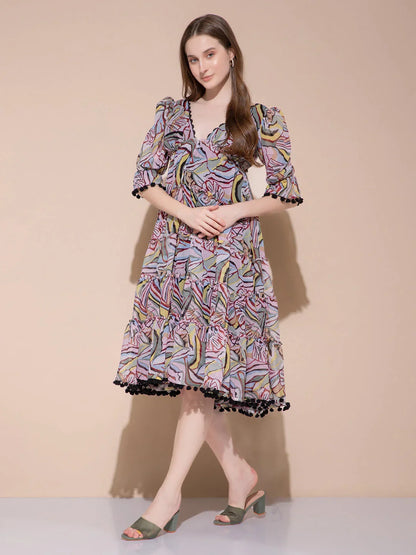 Woman in colorful abstract print midi dress with puff sleeves and black pompom trim posing against peach wall