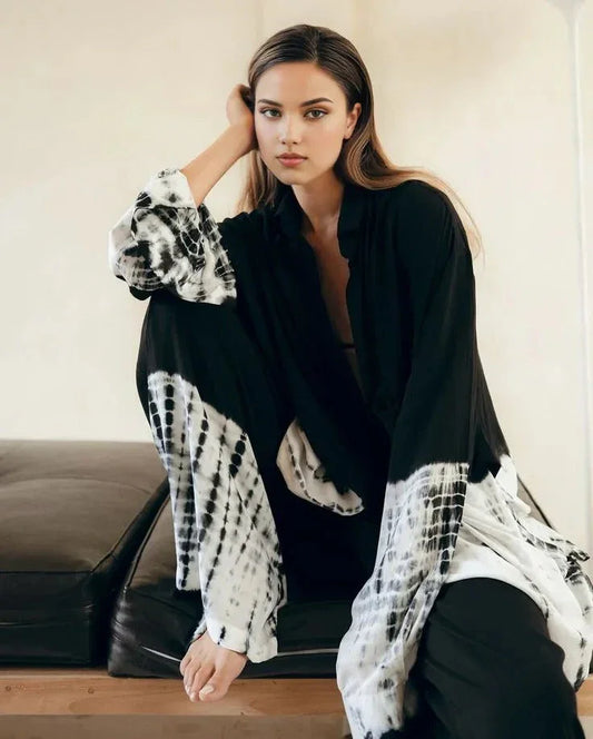 Woman wearing black and white tie-dye robe sitting on cushioned bench in minimalist room