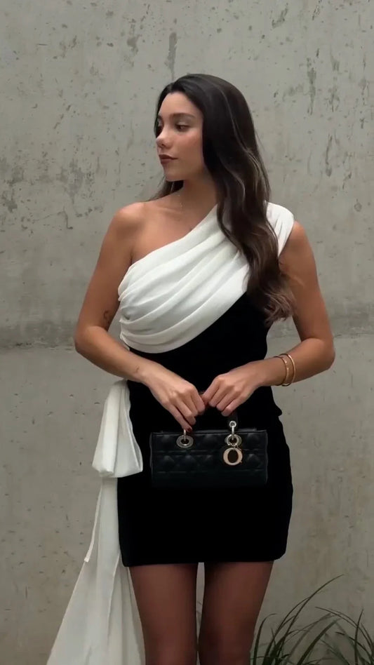 Woman wearing black and white one-shoulder short dress holding black handbag against concrete wall