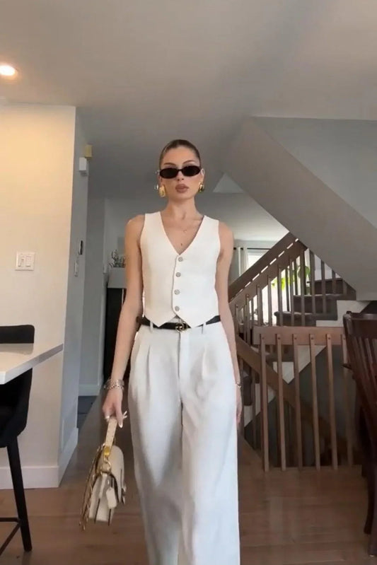 Stylish woman in white vest and wide-leg pants holding beige handbag in modern home interior