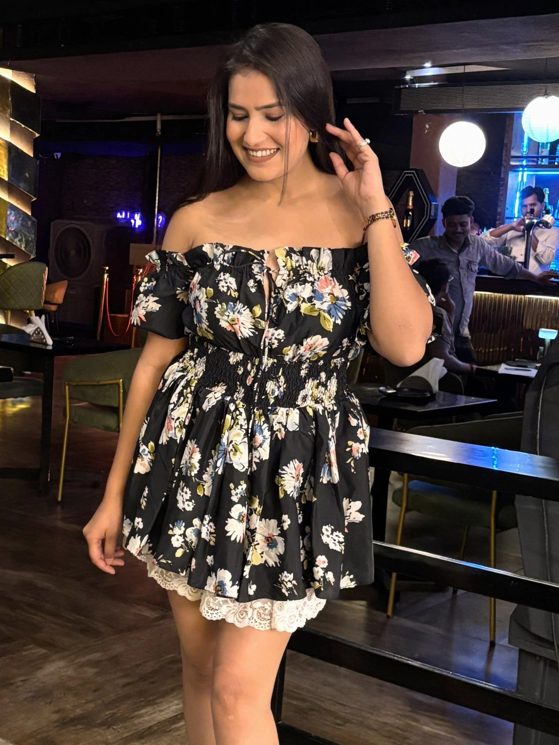 Smiling woman in a black floral off-shoulder dress with lace trim in a dimly lit bar setting