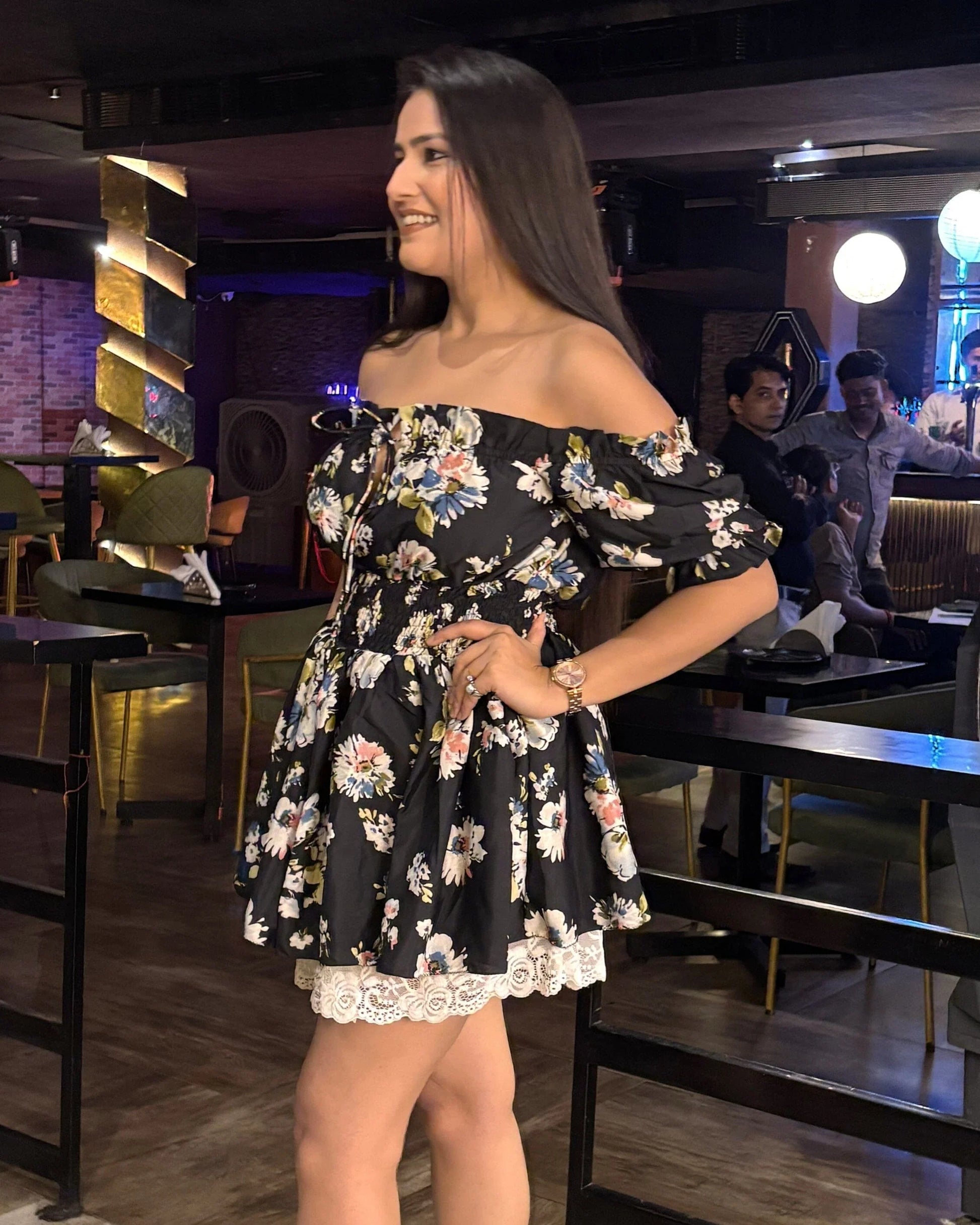 Woman in a black floral off-shoulder dress standing inside a dimly lit restaurant or lounge