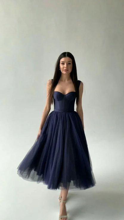 Woman in elegant navy blue tulle midi dress with fitted bodice and strapped heels against plain background